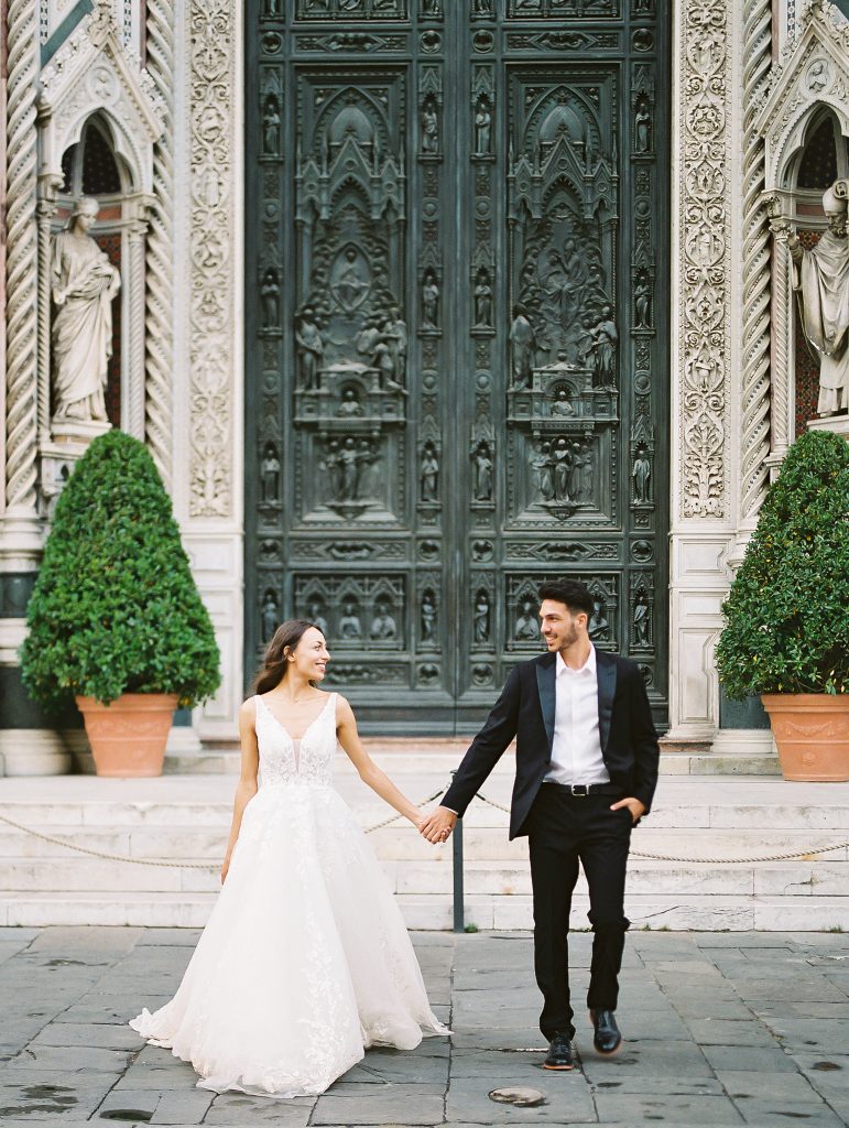 A wedding couple standing in front of the Cathedral of Santa Maria del Fiore also known as the Florence Duomo, the bride is wearing a lace dress with thick straps and the groom is wearing a black suit and white shirt with no tie, they are walking towards the camera but looking at each other smiling, image photographed by Kelley Williams a Florence Italy wedding photographer