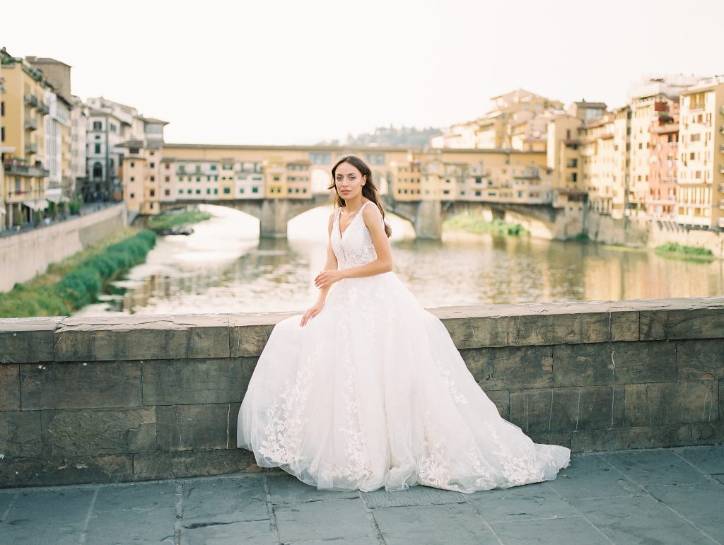 A close up portrait of a bride in Florence Italy taking a break sitting on the edge of a bridge with Ponte Vecchio in the background, she has beautiful blue eyes, image taken by Kelley Williams a Florence Italy wedding photographer