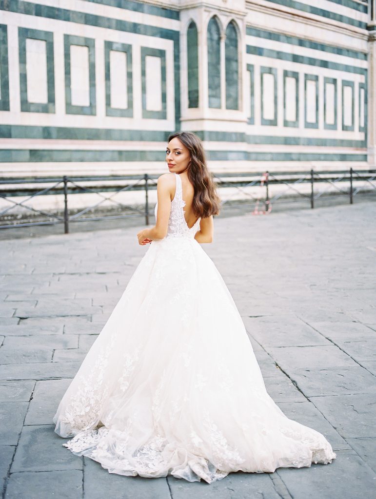 Portrait of a bride standing in front of the Cathedral of Santa Maria del Fiore also known as the Florence Duomo, the bride is wearing a lace dress with thick straps she is looking at the camera back over her shoulder, image photographed by Kelley Williams a Florence Italy wedding photographer