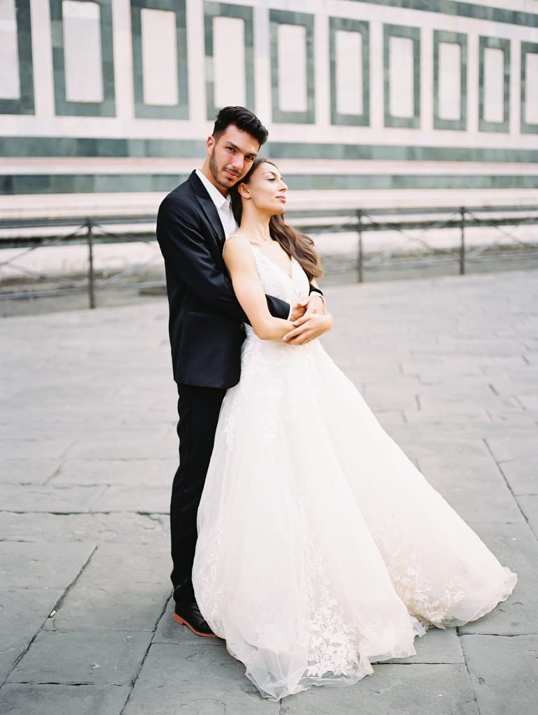 A wedding couple standing in front of the Cathedral of Santa Maria del Fiore also known as the Florence Duomo, the bride is wearing a lace dress with thick straps and the groom is wearing a black suit and white shirt with no tie, they are in an embrace and both of them are looking at the camera, image photographed by Kelley Williams a Florence Italy wedding photographer