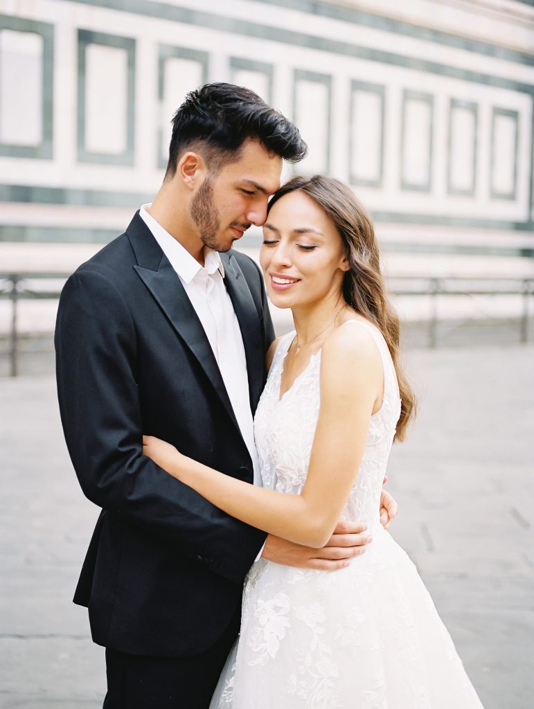 A wedding couple standing in front of the Cathedral of Santa Maria del Fiore also known as the Florence Duomo, the bride is wearing a lace dress with thick straps and the groom is wearing a black suit and white shirt with no tie, they are in an embrace not looking at the camera, image photographed by Kelley Williams a Florence Italy wedding photographer