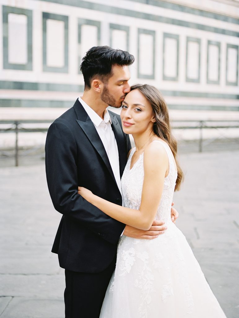 A wedding couple standing in front of the Cathedral of Santa Maria del Fiore also known as the Florence Duomo, the bride is wearing a lace dress with thick straps and the groom is wearing a black suit and white shirt with no tie, they are in an embrace and both of them are looking at the camera, image photographed by Kelley Williams a Florence Italy wedding photographer