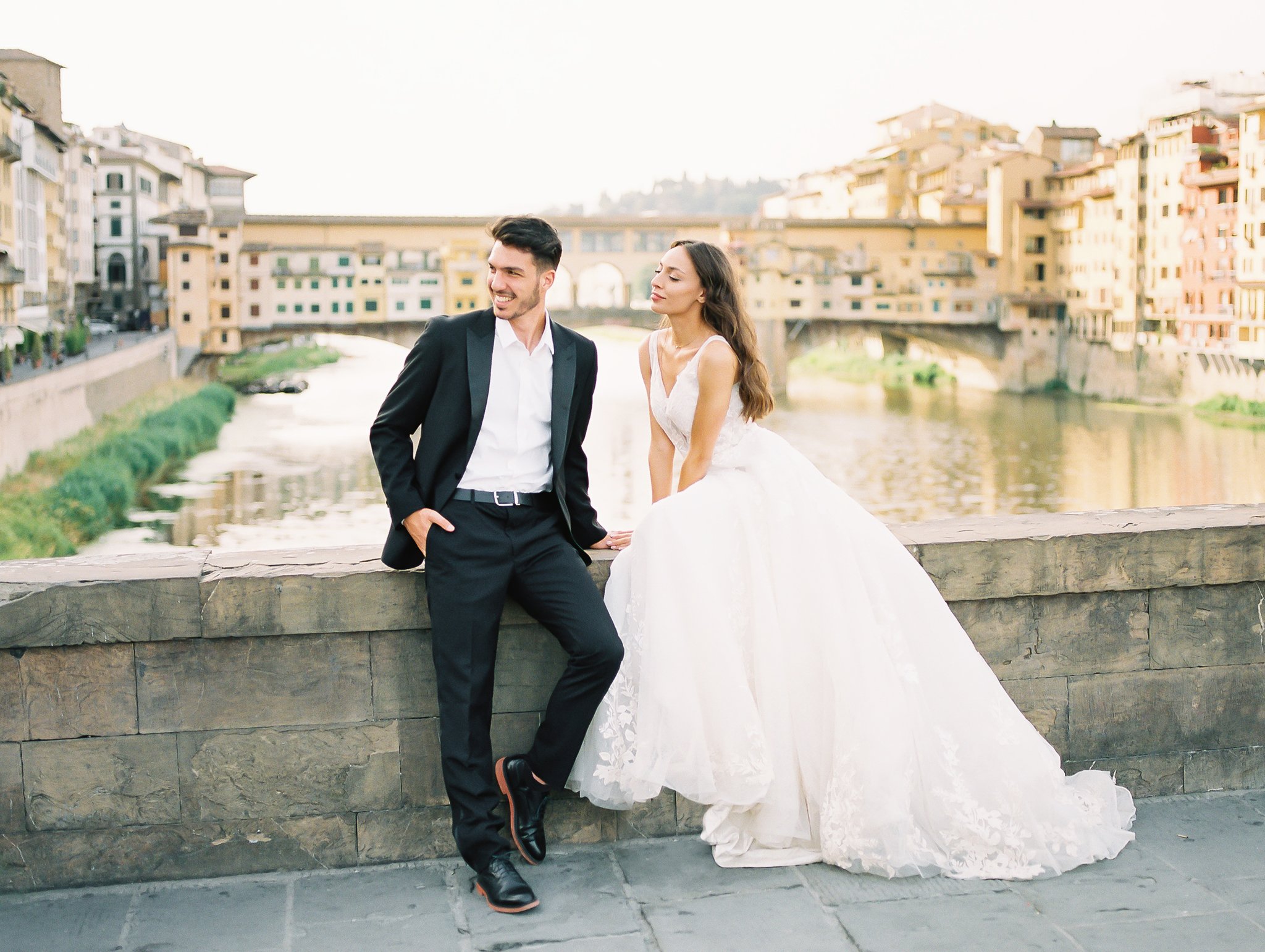 A wedding couple in Florence Italy taking a break sitting on the edge of a bridge with Ponte Vecchio in the background, image taken by Kelley Williams a Florence Italy wedding photographer