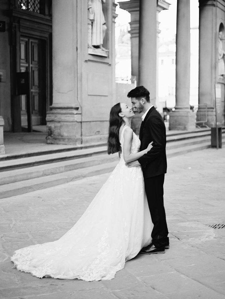 A wedding couple walking through the streets of Florence Italy, the bride is wearing a lace dress with thick straps and the groom is wearing a black suit and white shirt with no tie, they are holding hands and walking away from the camera, image photographed by Kelley Williams a Florence Italy wedding photographer