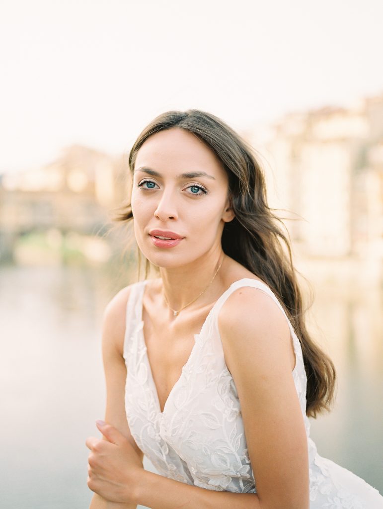 A close up portrait of a bride in Florence Italy taking a break sitting on the edge of a bridge with Ponte Vecchio in the background, she has beautiful blue eyes, image taken by Kelley Williams a Florence Italy wedding photographer