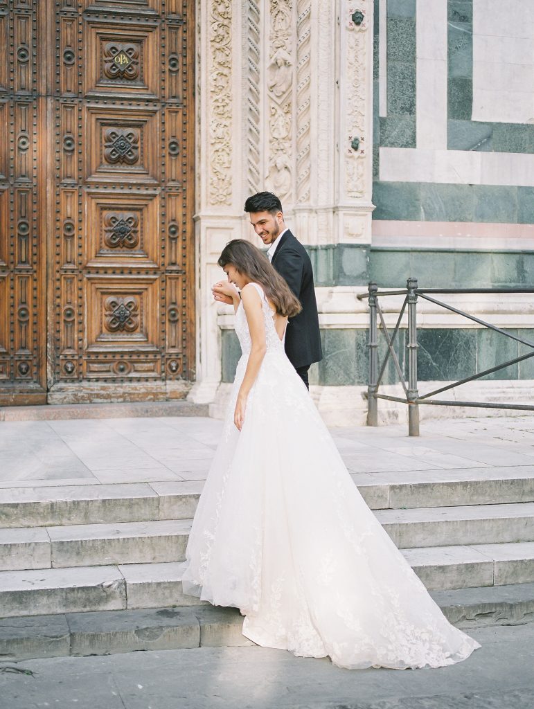 A wedding couple standing in front of the Cathedral of Santa Maria del Fiore also known as the Florence Duomo, the bride is wearing a lace dress with thick straps and the groom is wearing a black suit and white shirt with no tie, they are walking up the stairs to enter, image photographed by Kelley Williams a Florence Italy wedding photographer