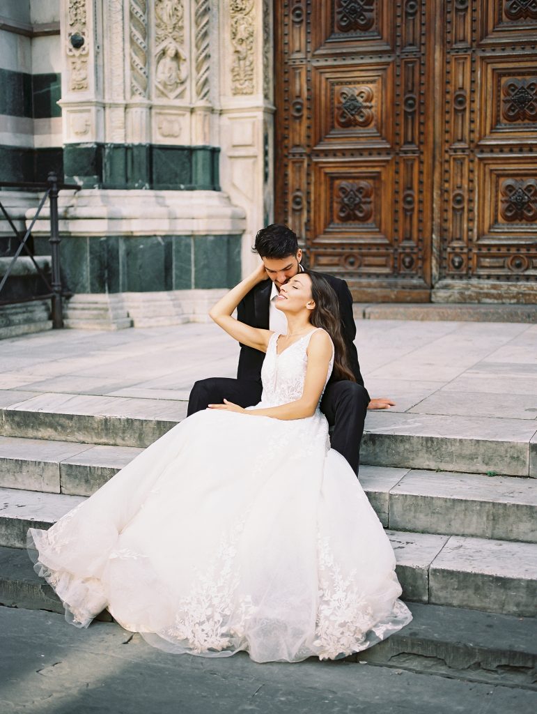 A wedding couple sitting on the steps in front of the Cathedral of Santa Maria del Fiore also known as the Florence Duomo, the bride is wearing a lace dress with thick straps and the groom is wearing a black suit and white shirt with no tie, they are in an embrace and kissing, image photographed by Kelley Williams a Florence Italy wedding photographer