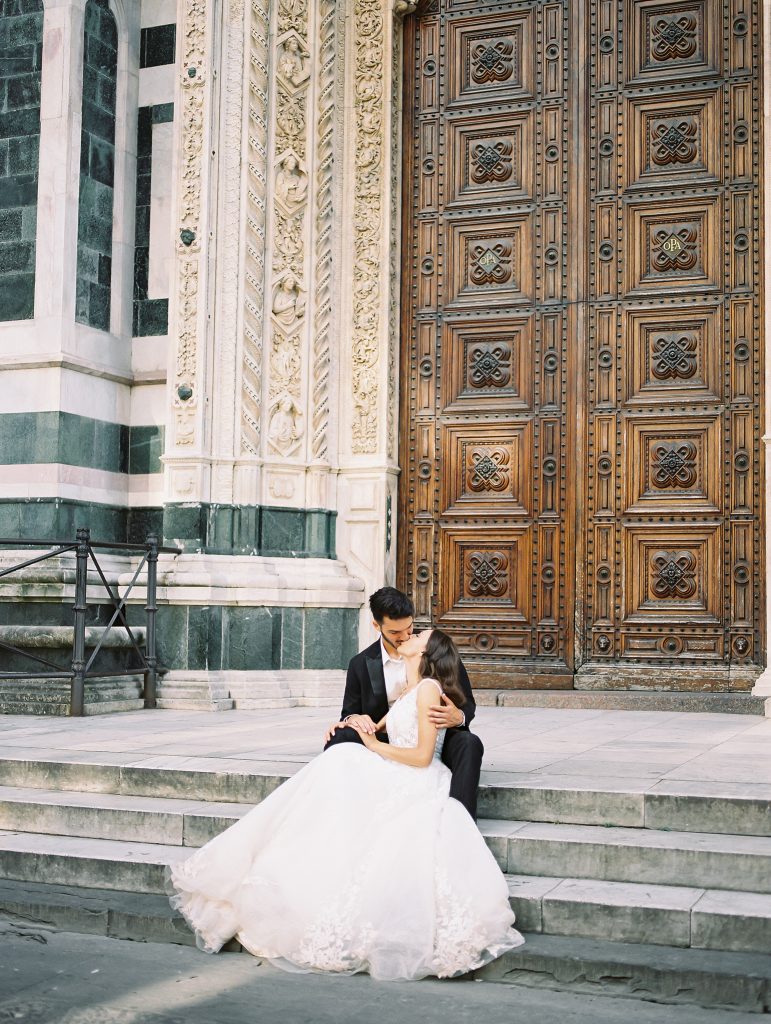 A wedding couple sitting on the steps in front of the Cathedral of Santa Maria del Fiore also known as the Florence Duomo, the bride is wearing a lace dress with thick straps and the groom is wearing a black suit and white shirt with no tie, they are in an embrace and kissing, image photographed by Kelley Williams a Florence Italy wedding photographer