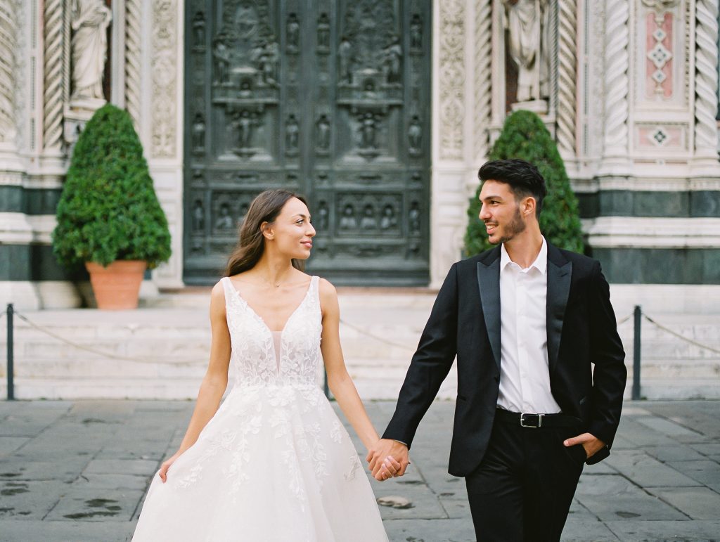A wedding couple standing in front of the Cathedral of Santa Maria del Fiore also known as the Florence Duomo, the bride is wearing a lace dress with thick straps and the groom is wearing a black suit and white shirt with no tie, they are walking towards the camera but looking at each other smiling, image photographed by Kelley Williams a Florence Italy wedding photographer