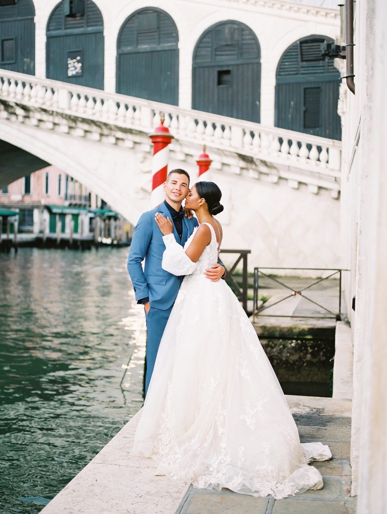 A wedding couple standing on the edge of the water in front of the famous Rialto Bridge in Venice Italy, groom is wearing a light blue suit with a navy shirt and bow tie the bride is wearing a lace wedding dress with sleeves and they are looking at each other after eloping image photographed by Kelley Williams Photography a Venice Italy wedding photographer. 