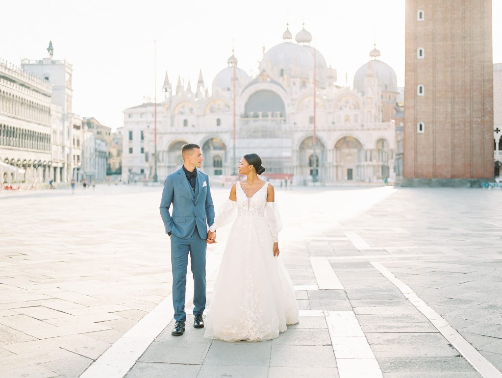 A couple in front of the grand cathedral in San Marcos square groom is wearing a light blue 
 suit with a navy shirt and bow tie the bride is wearing a lace wedding dress with sleeves and they are looking at each other after eloping image photographed by Kelley Williams Photography a Venice Italy wedding photographer. 