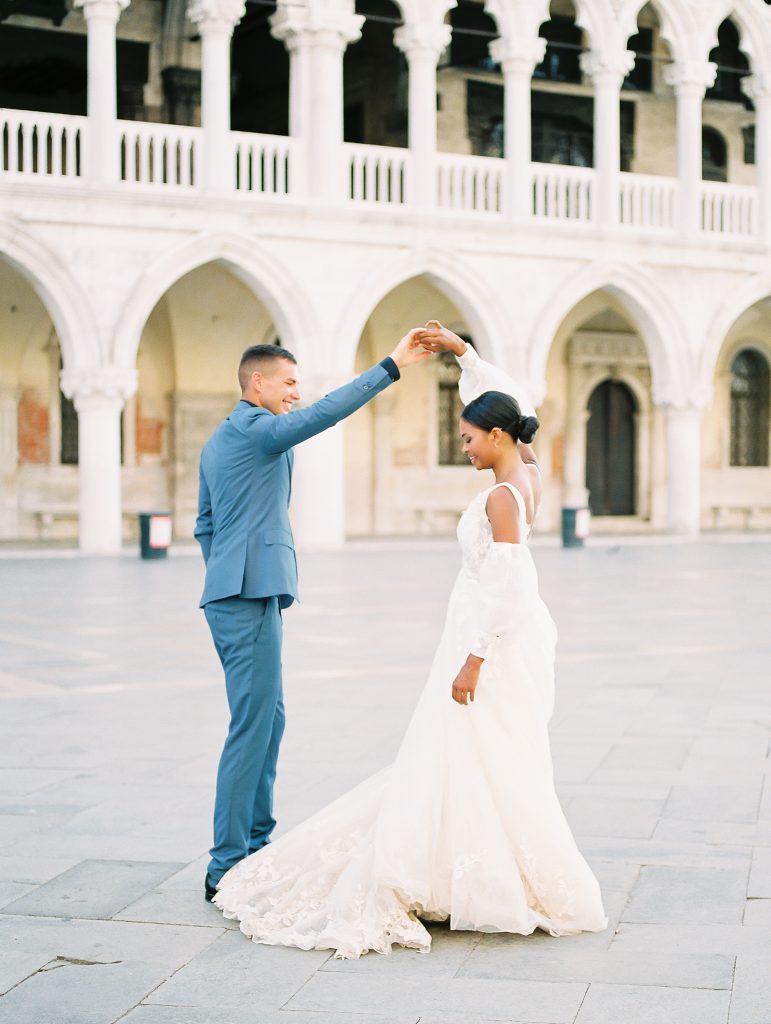 A wedding couple in front of a white building with arches and columns and the groom is giving the bride a twirl the groom is wearing a light blue suit with a navy shirt and bow tie the bride is wearing a lace wedding dress with sleeves and they are smiling the image photographed by Kelley Williams Photography a Venice Italy wedding photographer. 