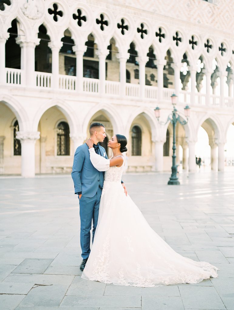 A wedding couple in front of a white building with arches and columns and the bride and groom are looking at each other in an embrace, the groom is wearing a light blue suit with a navy shirt and bow tie the bride is wearing a lace wedding dress with sleeves and they are smiling the image photographed by Kelley Williams Photography a Venice Italy wedding photographer. 