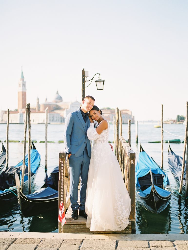 A wedding couple in an embrace on a deck ramp of gondolas groom is wearing a light blue suit with a navy shirt and bow tie the bride is wearing a lace wedding dress with sleeves image photographed by Kelley Williams Photography a Venice Italy wedding photographer. 