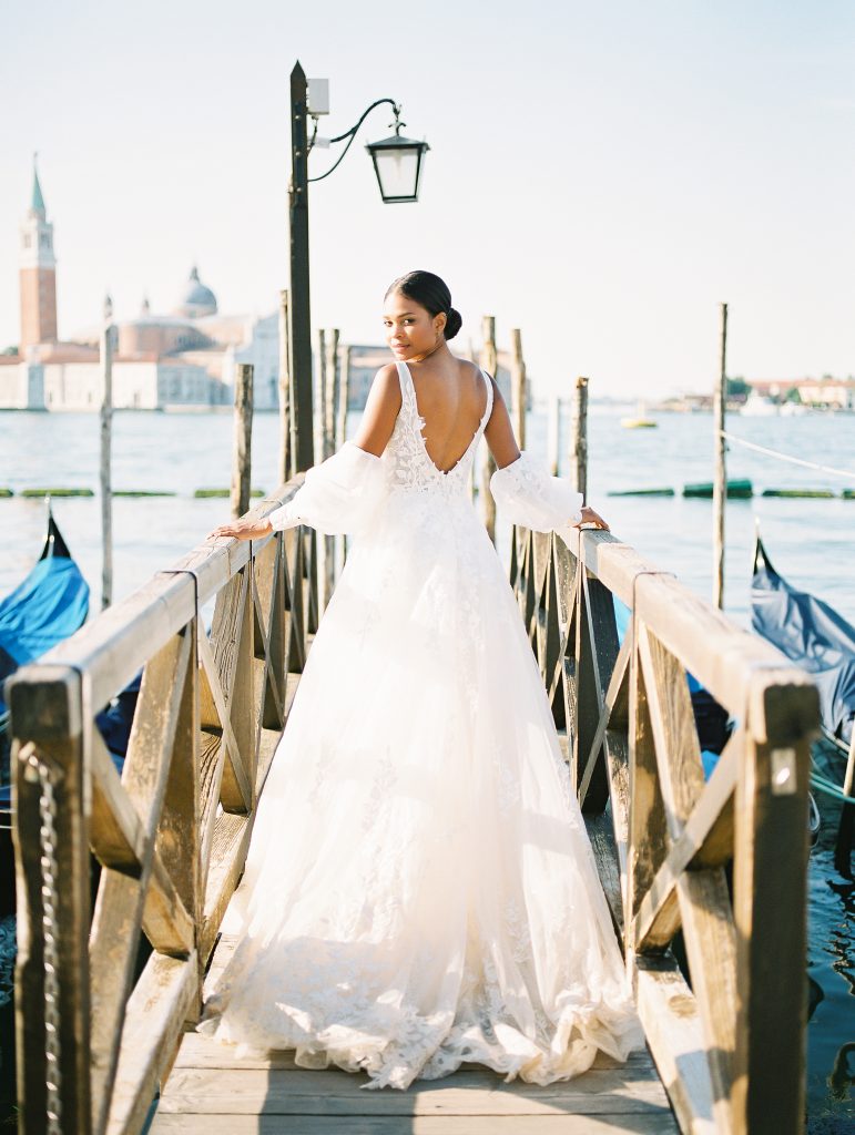 A bride looking over her shoulder at the camera wearing a lace wedding dress with sleeves, she is standing on a dock with gondolas around her the image photographed by Kelley Williams Photography a Venice Italy wedding photographer. 