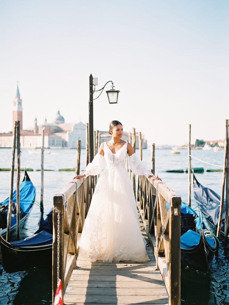 A bride facing the camera looking over to her right, wearing a lace wedding dress with sleeves, she is standing on a dock with gondolas around her the image photographed by Kelley Williams Photography a Venice Italy wedding photographer. 