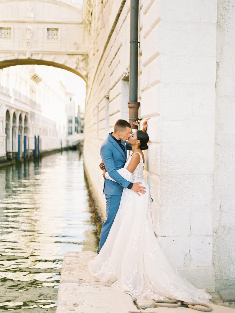 A couple near the edge of the water in Venice Italy the groom is wearing a light blue suit with a navy shirt and bow tie the bride is wearing a lace wedding dress with sleeves and they fully embraced and kissing after eloping image photographed by Kelley Williams Photography a Venice Italy wedding photographer. 
