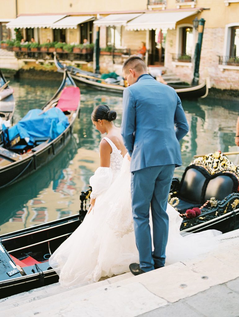 A wedding couple is walking down steps about to get into a black gondola, the bride is in front of the groom, and the groom is wearing a light blue suit with a navy shirt and bow tie the bride is wearing a lace wedding dress with sleeves image photographed by Kelley Williams Photography a Venice Italy wedding photographer. 