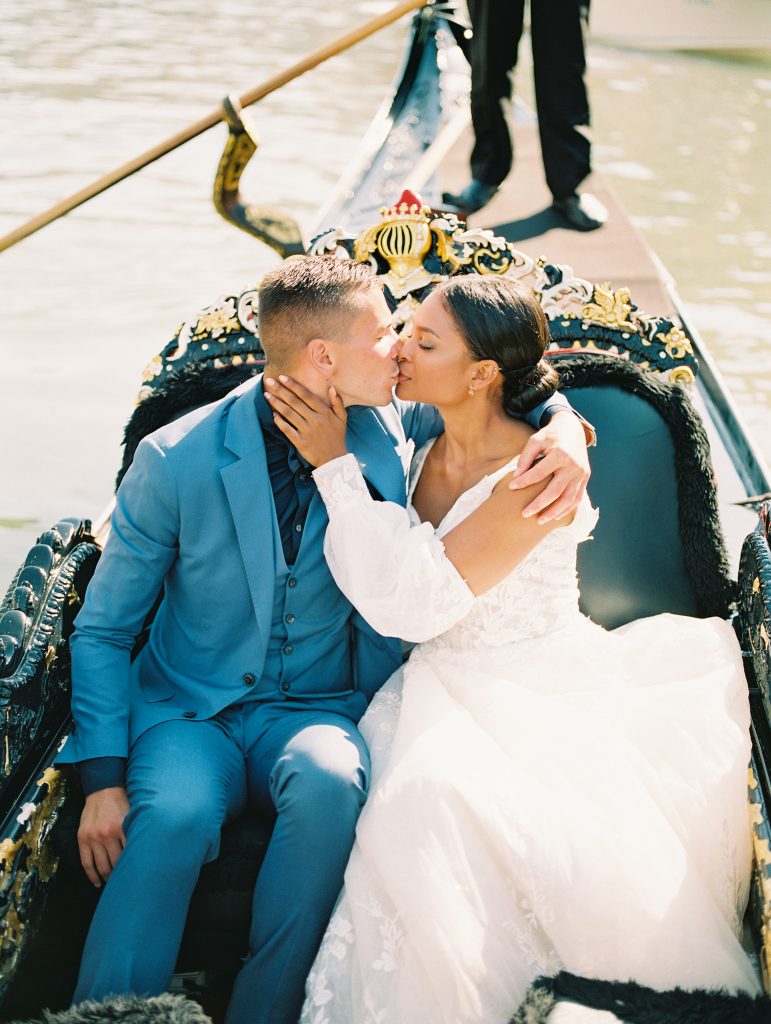 A wedding couple kissing while sitting in a black gondola, the bride is in front of the groom, and the groom is wearing a light blue suit with a navy shirt and bow tie the bride is wearing a lace wedding dress with sleeves image photographed by Kelley Williams Photography a Venice Italy wedding photographer. 