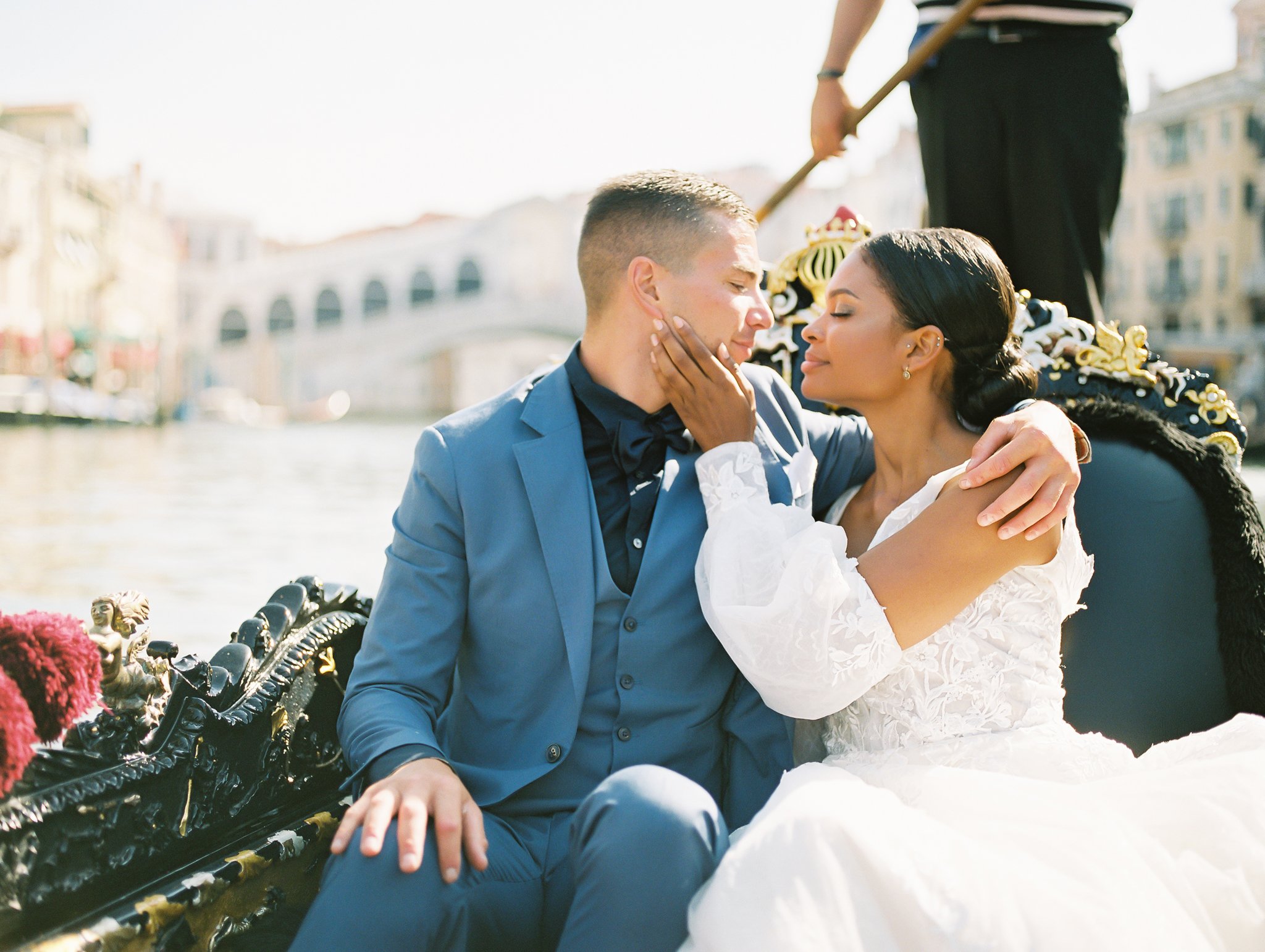 A wedding couple in gondola in one of the many venice italy canals the gondola is black with gold trimmings the groom is wearing a light blue suit with a navy shirt and bow tie the bride is wearing a lace wedding dress with sleeves and the bride is looking at the camera but the groom is leaning on the bride looking down image photographed by Kelley Williams Photography a Venice Italy wedding photographer.