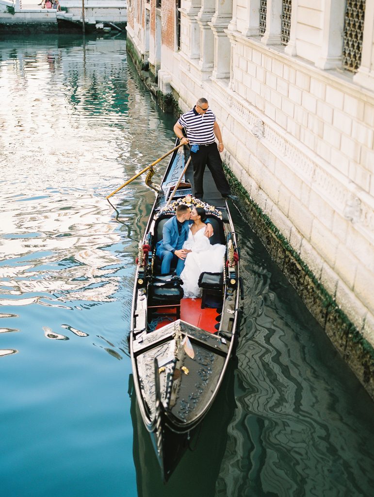 A wedding couple sharing a kiss while sitting inside of a gondola, the Gondola driver also known as a gondolier is rowing the boat along the canals in Venice Italy, image photographed by Kelley Williams Photography a Venice Italy wedding photographer. 