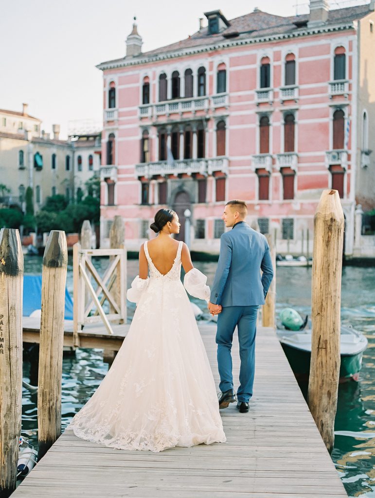 A couple in front of a pink building on the Venice Italy canal and they are walking away from the camera while standing on a dock groom is wearing a light blue suit with a navy shirt and bow tie the bride is wearing a lace wedding dress with sleeves and they are looking at each other after eloping image photographed by Kelley Williams Photography a Venice Italy wedding photographer. 
