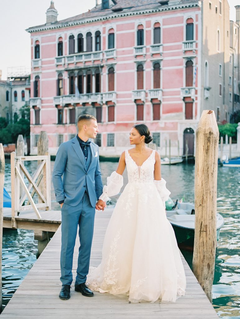 A couple in front of a pink building on the Venice Italy canal and they are walking towards the camera while standing on a dock groom is wearing a light blue suit with a navy shirt and bow tie the bride is wearing a lace wedding dress with sleeves and they are looking at each other after eloping image photographed by Kelley Williams Photography a Venice Italy wedding photographer. 