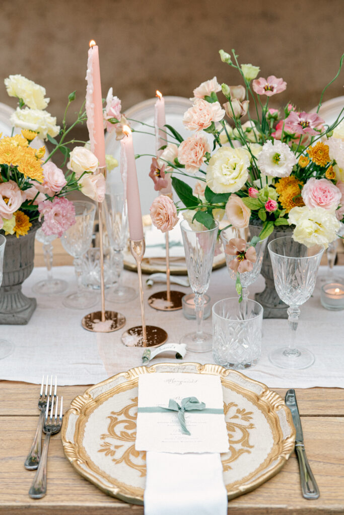 Close up of a table setting, gold chargers, lit pink candles and flowers with pink, peach, blush, and yellow, Abbazia San Pietro in Valle wedding, image taken by Kelley Williams a wedding photographer in Umbria Italy